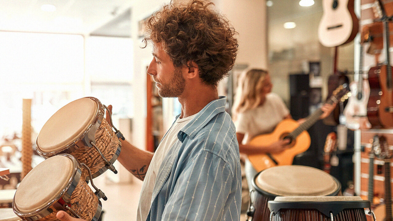 Persoon bekijkt slagwerkinstrumenten in een muziekwinkel, met gitaren en andere muziekinstrumenten op de achtergrond.