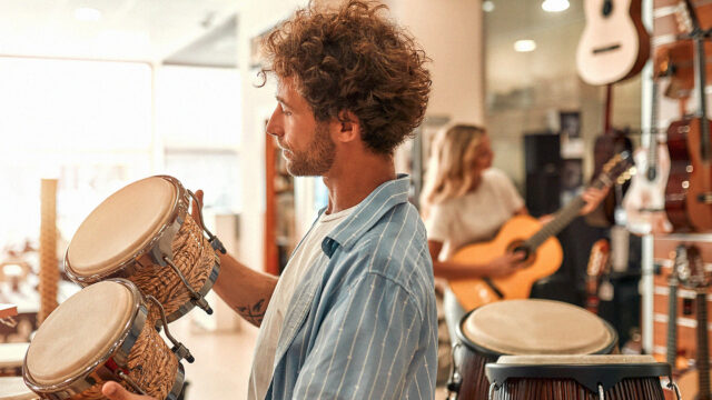 Persoon bekijkt slagwerkinstrumenten in een muziekwinkel, met gitaren en andere muziekinstrumenten op de achtergrond.