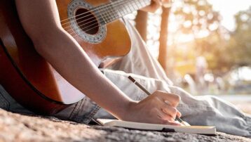 Een muzikant schrijft songteksten in een notitieboek terwijl ze gitaar speelt in de buitenlucht, met zonlicht op de achtergrond.