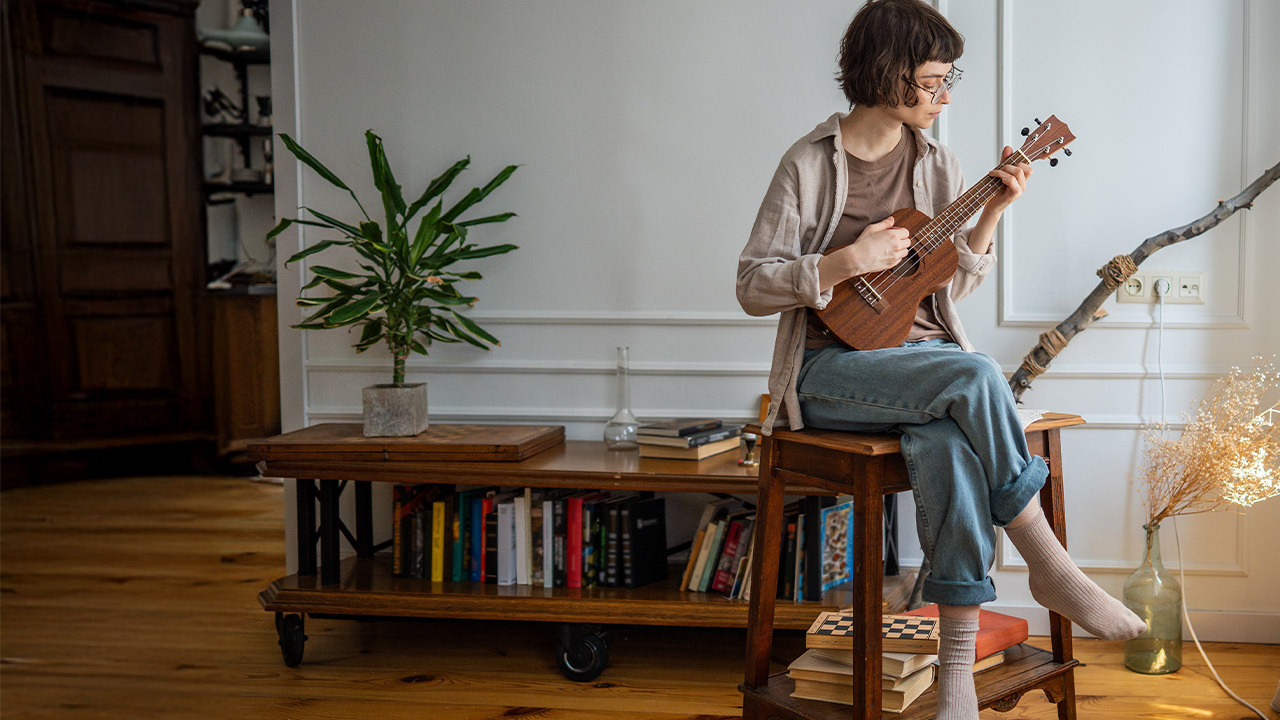 Persoon die binnenshuis ukelele speelt, als symbool voor de mentale en emotionele voordelen van het leren bespelen van een muziekinstrument