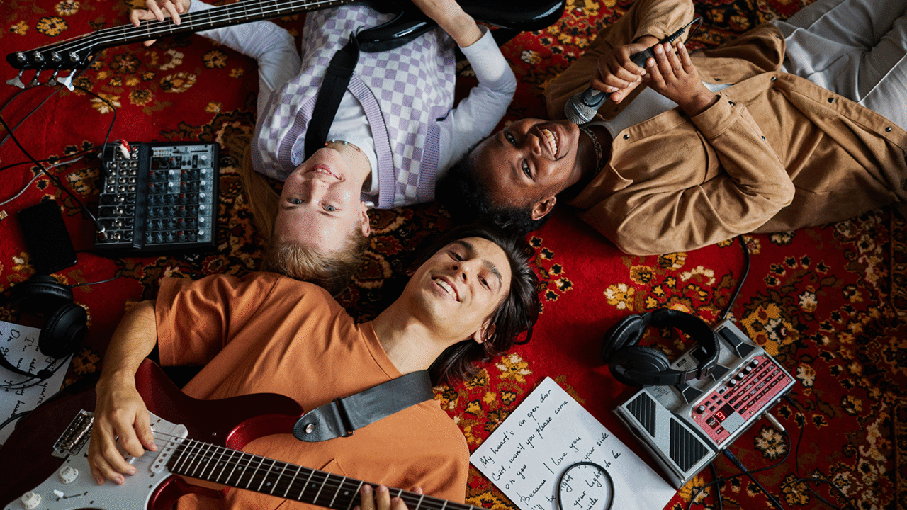 Groep jonge amateurmuzikanten die ontspannen op een tapijt liggen met gitaren, bas, microfoon en effectpedalen, terwijl ze samen muziek maken – amateurmuzikant zijn is nog steeds geweldig