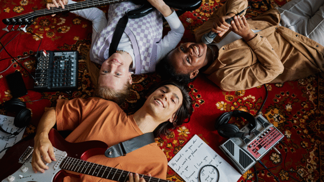 Groep jonge amateurmuzikanten die ontspannen op een tapijt liggen met gitaren, bas, microfoon en effectpedalen, terwijl ze samen muziek maken – amateurmuzikant zijn is nog steeds geweldig
