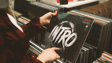 Iemand bladert door een collectie vinylplaten en houdt een platenhoes met het woord 'Intro' vast. De afbeelding staat symbool voor de meest iconische songintro's uit de muziekgeschiedenis.